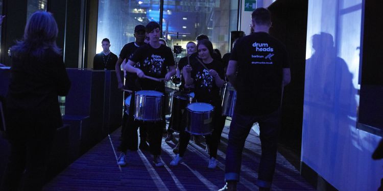A group of drummers perform in the Milton Court Foyers under low lighting