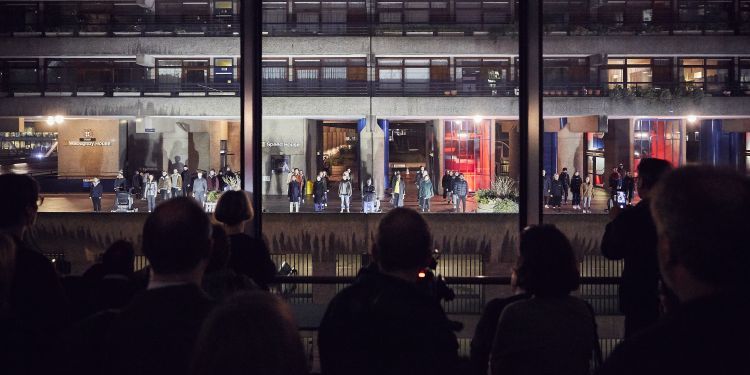 Performers stand illuminated on the Barbican highwalk, while silhouetted audience members look on from inside Milton Court