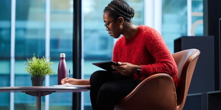 Woman seated on chair, with iPad in one hand and writing with the other. 