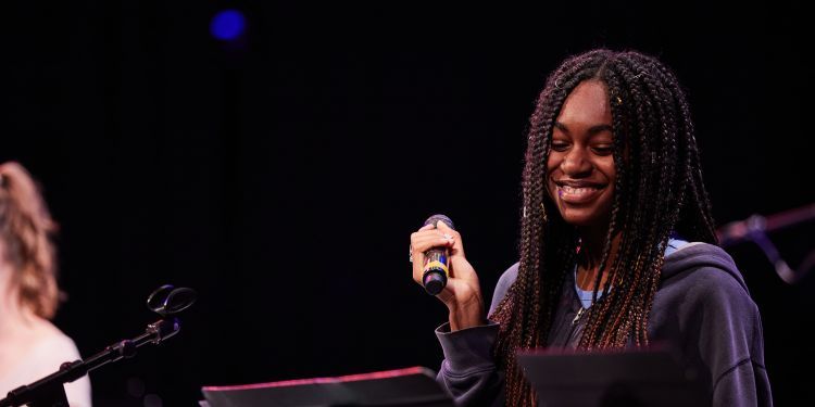 Woman on stage holding microphone smiling and looking down at music on stand