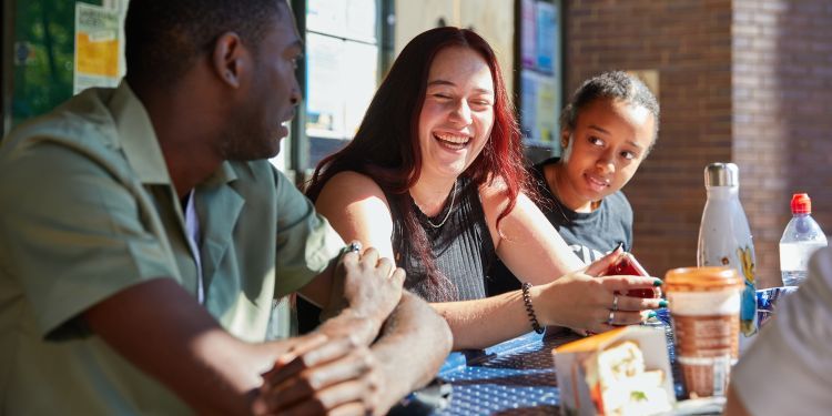 Students sitting at a bench table on Lakeside terrace, laughing while having lunch