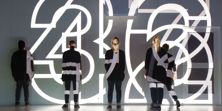 Large white neon numbers projected to the back of a stage over performers standing facing the back