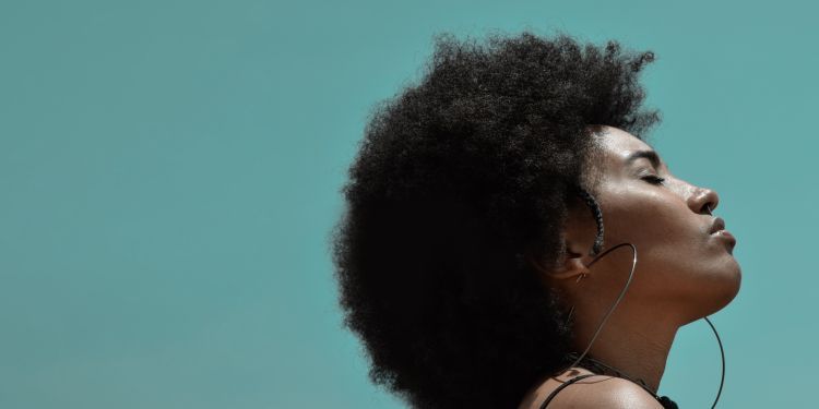 Close up of young black woman with afro hair raising their head and closing their eyes in the sunshine, against a green background
