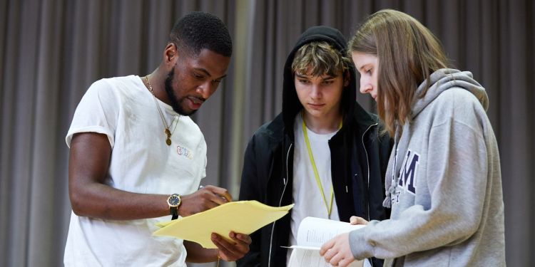 A man in a white t-shirt stands holding a piece of paper. To his left is a boy wearing a black hoodie and next to him is a girl with a grey hoodie. 