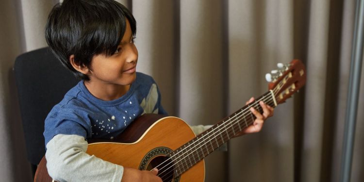 A boy with black hair and long sleeved blue and grey top smiles away from the camera as he plays the guitar