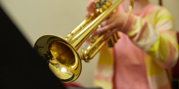 A close up image of a girl wearing a pink and yellow striped cardigan playing the trombone. The girls face is out of shot. 