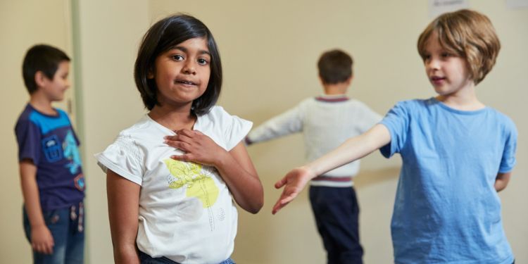 A girl with black hair and a white t-shirt looks at the camera, her hand touching her chest. A boy in blue stands next to her with his arm out. Two other boys can be seen in the distance.