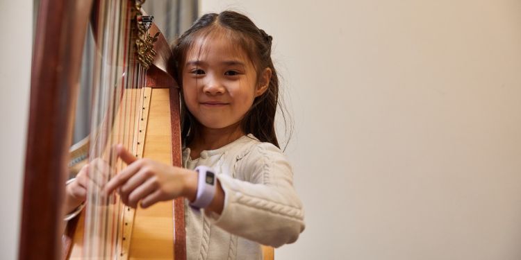 A girl with black hair and wearing a white knitted dress smiles at the camera whilst playing a harp