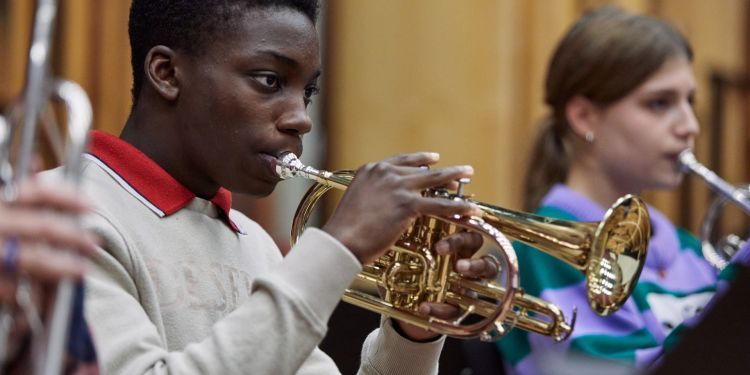 A boy wearing a white jumper, with a red collar sticking out of it, plays the trumpet 