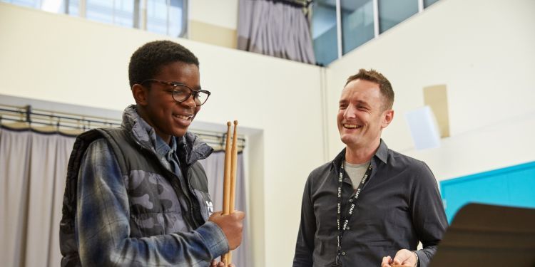 A boy in a blue checked t-shirt and camouflaged gilet holds a pair of drum sticks. He is stood next to a man in a grey shirt. Both are smiling.