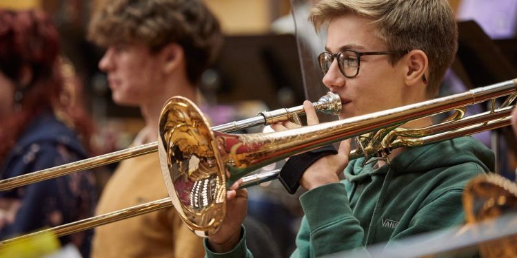 A boy with blonde hair and wearing a green jumper is sitting down playing a trombone
