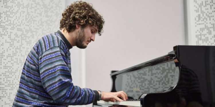 A boy with brown curly hair and a striped blue and black jumper sits playing the piano 