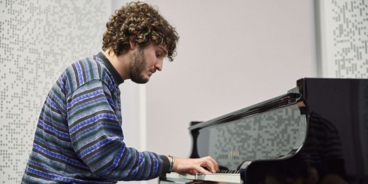 A boy with brown curly hair and a striped blue and black jumper sits playing the piano