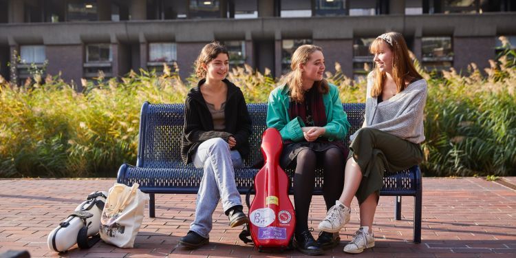 Three girls sit on a bench. From left to right: One wears a black cardigan, one wears a green jacket and the other wears a grey jumper. A red instrument case stands propped up against the bench in the middle of them.