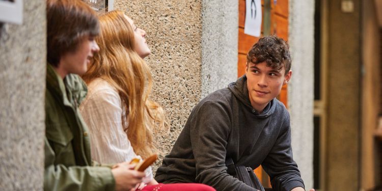 A boy wearing a grey jumper sits and faces a girl in a white shirt and a boy in a green jacket. He has a smile on his face. 