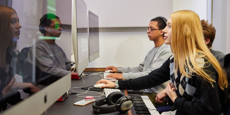 A girl with blonde hair and wearing a black jumper, with blue and beige  triangles, sits facing a large iMac