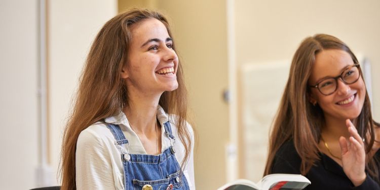 A girl with brown hair and wearing blue dungarees and a white shirt holds out a book. She has a big smile across her face. 