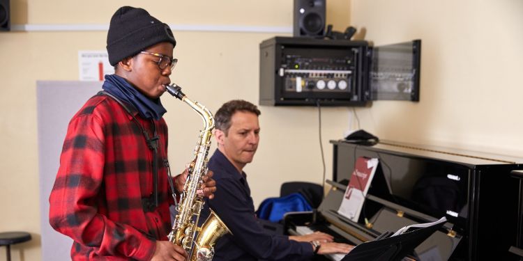 A boy with a black beanie hat, wearing a red and black checked shirt plays the saxophone. A man in a blue shirt plays the piano next to him.