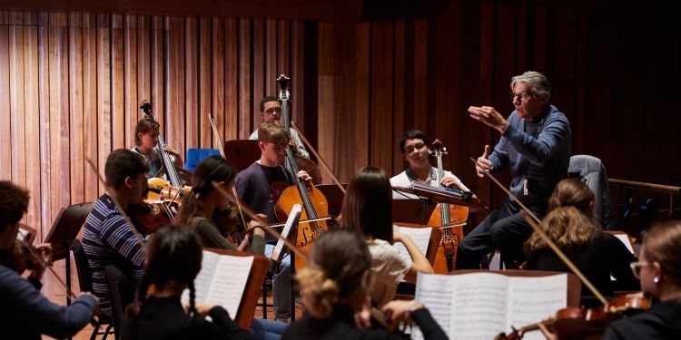 A man with grey hair and a blue jumper conducts an orchestra