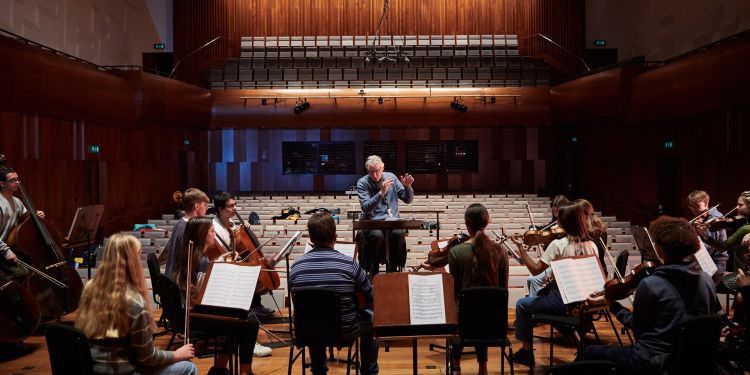 A man with grey hair and a blue jumper conducts an orchestra