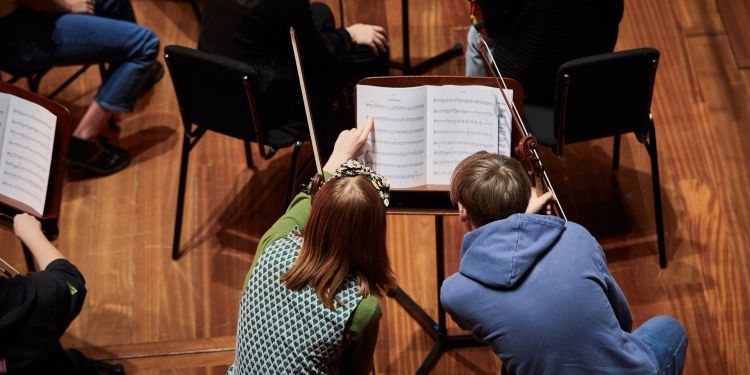 From left to right: A girl wearing a lime green long sleeve top with a spotted blue slip sits next to a boy in a blue jumper. They look at a music book together.