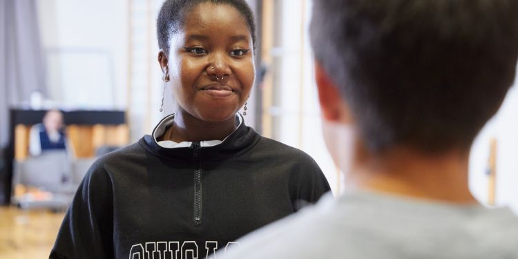 A girl with black hair tied up in a bun and wearing a black jumper smiles as she looks at the person stood in front of her
