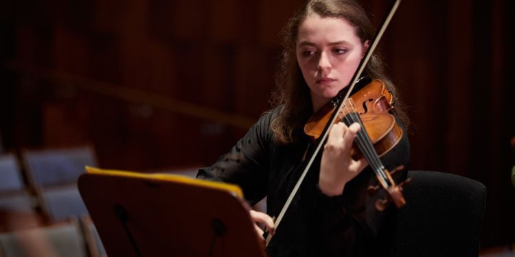 A girl with brown hair and wearing a black long sleeved top plays the violin. 
