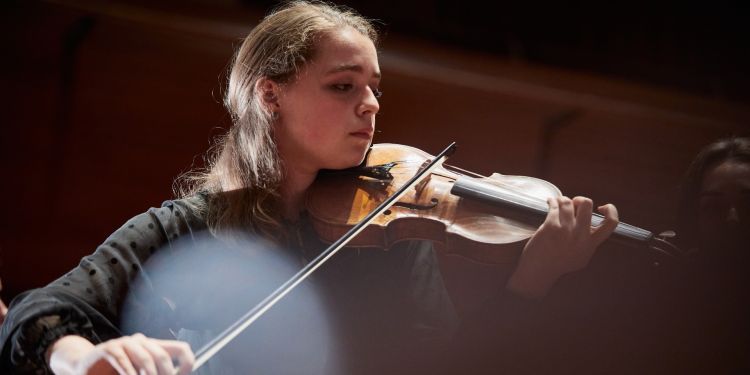 A girl with brown hair and wearing a black long sleeved top plays the violin. 