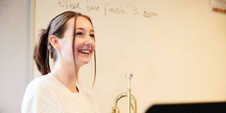 A girl in a white jumper is holding a trumpet. She has a big smile on her face