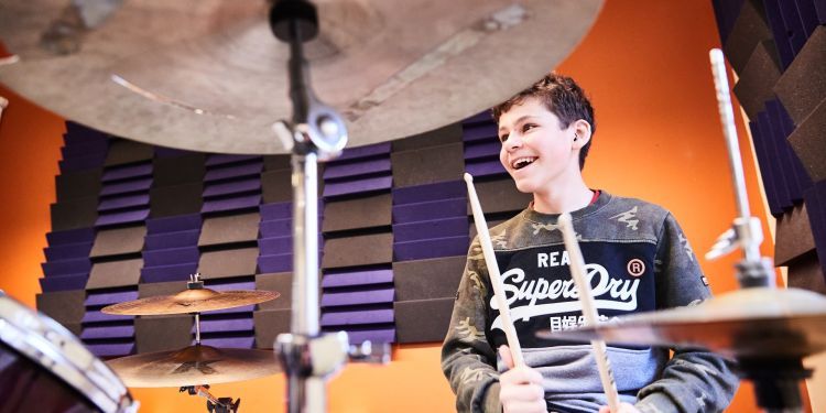 A boy wearing a Superdry jumper is playing the drums. He has a big smile on his face