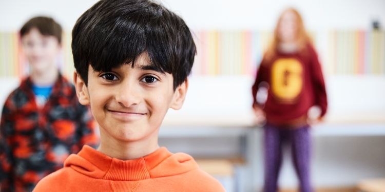 A young boy with black hair and wearing a bright orange jumper smiles at the camera