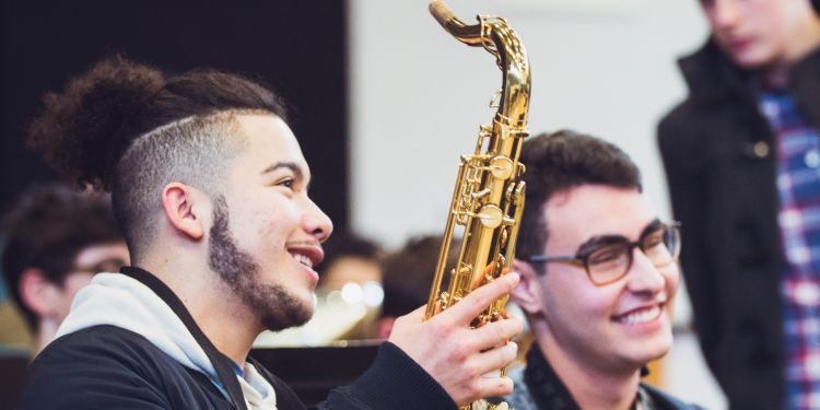 A boy in a blue jacket holds up a saxophone. He sits next to another boy. Both are smiling