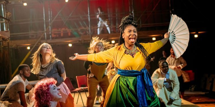 Actors dance on stage, one in yellow and green dress holding arms outstretched with a fan in one hand