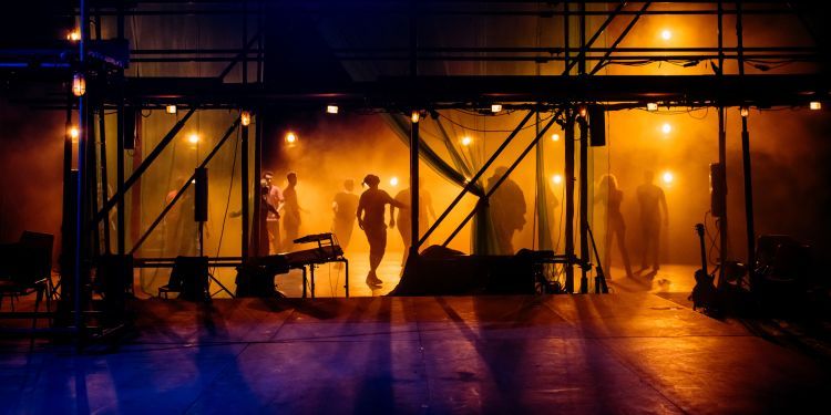 Actors silhouetted by orange light at the back of the stage as they dance under scaffolding