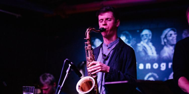 A man wearing a dark blazer and t-shirt plays a saxophone on-stage. There are projections in the background