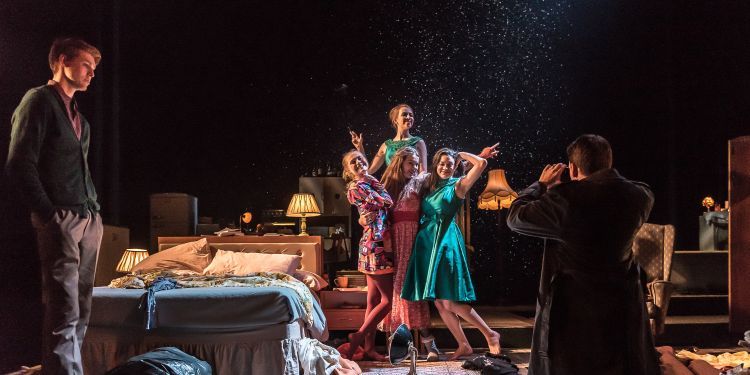 Actors on stage in a bedroom set, posing for a photo while artificial snow falls around them