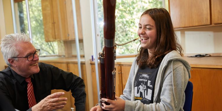 A girl with brown hair plays the oboe, she has a big smile on her face. A man in a black shirt and red tie crouches down next to her, he is also smiling. 