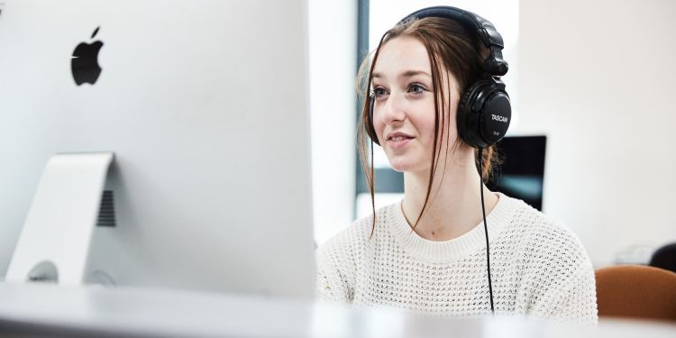 A girl with brown hair and wearing a white jumper sits in front of a big Apple Mac. Her hair is tied up and she is wearing a pair of black headphones.