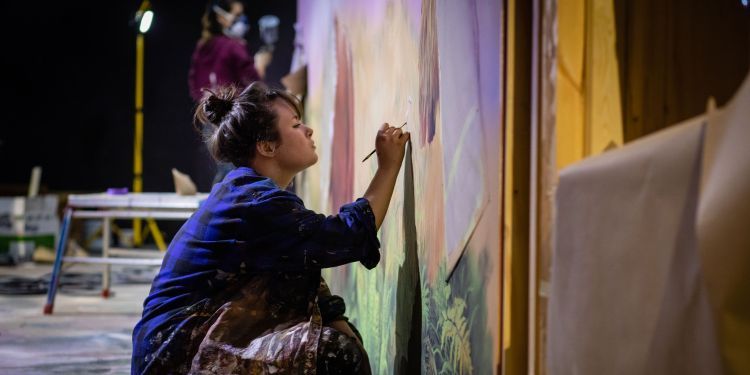 Student sits on the floor to paint a piece of the set for Beasts of London