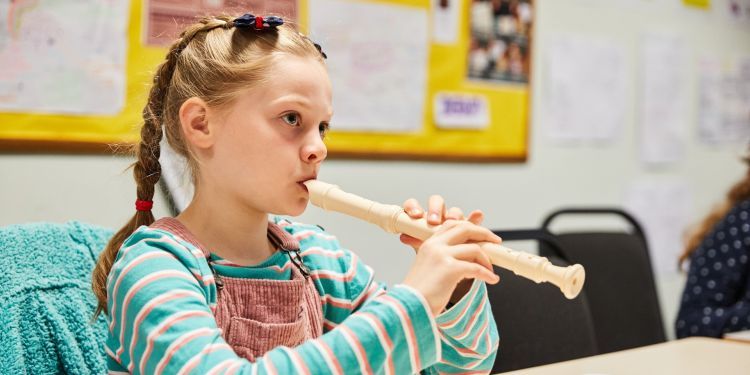 A young girl sits at a desk and plays a flute. She has blonde hair which she wears in two plaits 