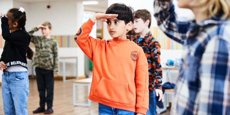 A boy with black hair wearing an orange jumper has his hand across his forehead