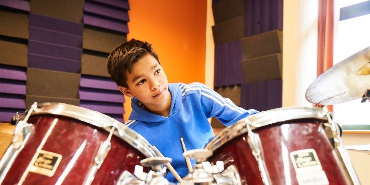 A boy with black hair and wearing a blue hoody sits behind a red drum kit