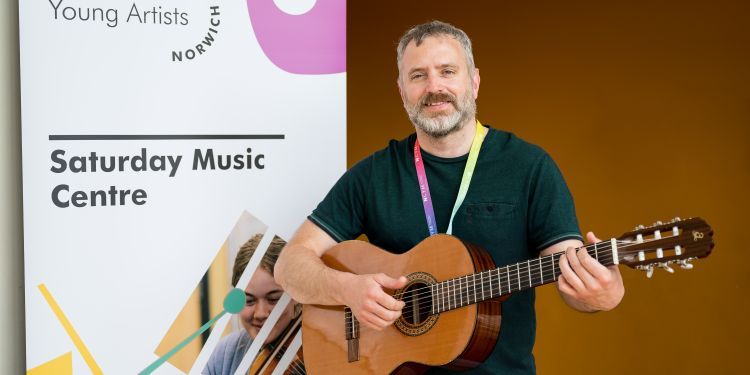 Guitar tutor in black t shirt holding guitar next to pop banner for GYA Norwich