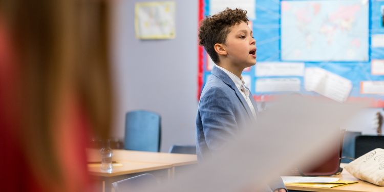 Boy in blue jacket singing with blue notice board behind him, blurred side image in front of student with long straight hair holding music