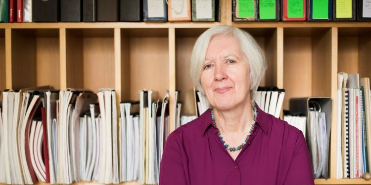 Judith Weir wears a purple shirt and stands infront of a bookcase