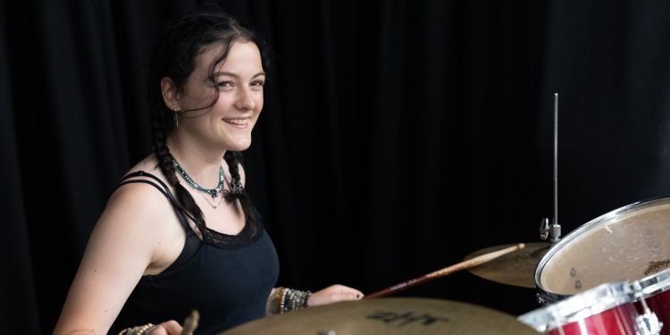 A girl with brown hair sits behind a red drum kit and smiles