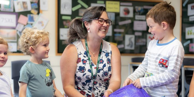 A female teacher smiles at the boy on her left. They are holding a purple sparkly cloth in front of them