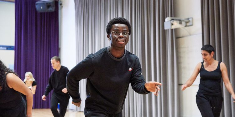 A boy smiles in class. He is wearing a black jumper and grey joggers