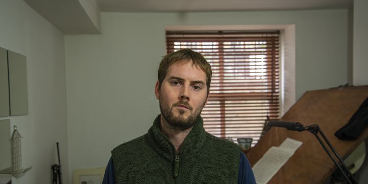 Composer Oliver Leith standing in room looking at the camera with music score on an easel behind him