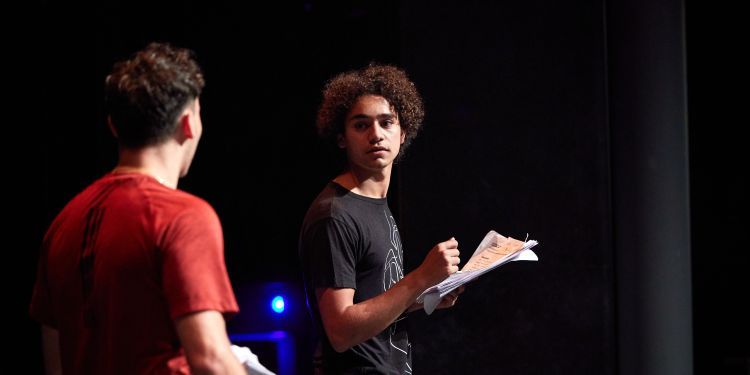 Two men on a stage turning towards each other, both are holding a script